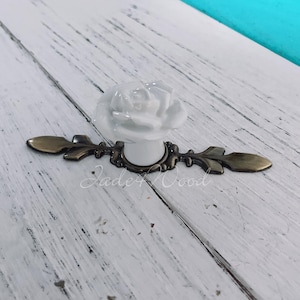 Könnte beinhalten: Weißer Porzellan-Rosenblüten-Schrankknopf mit Blattgold-Design. Der Knopf ist an einer Metallbasis mit einer Schraube befestigt.