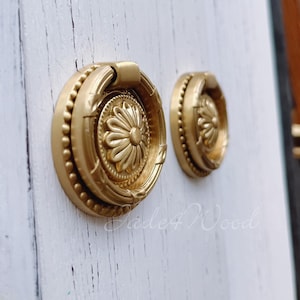 Pode incluir: Dois botões de armário dourados com um design floral decorativo e um puxador de anel. Os botões têm uma borda de contas.