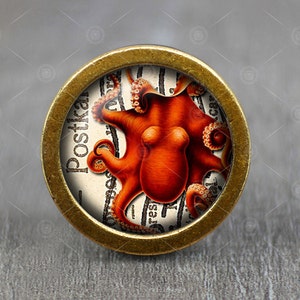 Puede incluir: Un pomo redondo de color latón con una imagen vintage de un pulpo naranja sobre un fondo blanco. El pulpo está rodeado de texto en un idioma extranjero. El pomo es perfecto para agregar un toque único a los muebles o armarios.