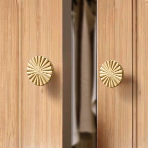 May include: Two gold-toned cabinet knobs with a sunburst design. The knobs are mounted on a wooden surface.