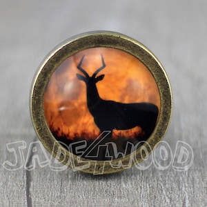 May include: A round, bronze-coloured cabinet knob with a glass insert featuring a silhouette of a deer against an orange sunset background.