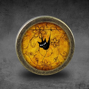 Könnte beinhalten: Ein runder, messingfarbener Knopf mit einer schwarzen Katzensilhouette, die an einer Linie gegen einen gelben Hintergrund mit einem floralen Muster hängt.