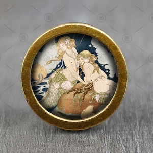 May include: A brass knob with a round, gold-colored frame and a glass insert featuring a vintage illustration of two mermaids with flowing hair and tails, surrounded by bubbles and seaweed.