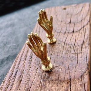 May include: Two gold-toned metal hands with raised fingers mounted on a rustic wooden board. The hands are positioned as if in prayer.