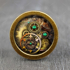 Pode incluir: Um pingente redondo de cor latão com um design steampunk. O pingente apresenta um mostrador de relógio com algarismos romanos e engrenagens, acentuado com gemas verdes.