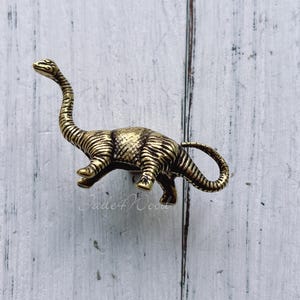 Op de afbeelding: Een gouden dinosaurusvormige kastknop of ladegreep. De dinosaurus staat met zijn staart om zijn lichaam gekruld.