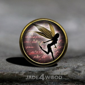May include: A round, gold-toned metal charm with a glass cabochon featuring a silhouette of a fairy with wings against a pink and white background with handwritten text.