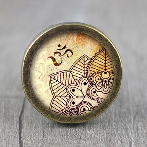 May include: A round, brass-colored knob with a vintage-style design featuring an Om symbol and a mandala pattern in shades of brown and cream.