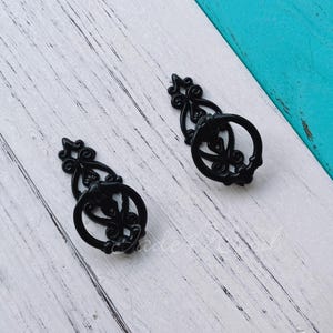 May include: Two black decorative drawer pulls with a circular handle and ornate detailing. The pulls are made of metal and have a vintage or antique style. They are set against a white and turquoise background.