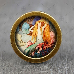 May include: A round brass knob with a colorful image of a mermaid with long flowing red hair, sitting on a coral reef. The mermaid is wearing a green tail and has a blue background.