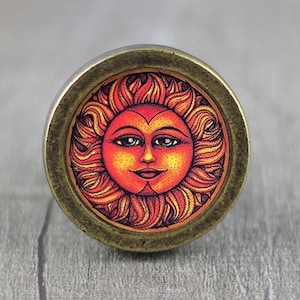 Puede incluir: Un pomo redondo de color latón con una imagen colorida de un sol sonriente con un centro en forma de corazón. El sol tiene una cara roja con rayos amarillos y naranjas.
