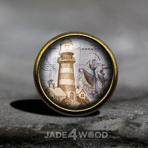 Könnte beinhalten: Ein runder, messingfarbener Knopf mit weißem Hintergrund, der eine handgezeichnete Illustration eines Leuchtturms und eines Schiffes zeigt. Der Leuchtturm ist mit Weiß und Braun gestreift, und das Schiff hat braune Segel und einen weißen Rumpf. Der Hintergrund ist eine Karte mit handschriftlichem Text.