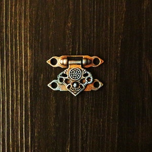 May include: A silver and gold metal clasp with a decorative design. The clasp has two loops on either side and a central decorative piece.