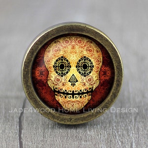 Puede incluir: Pomo de armario redondo con un diseño de calavera del Día de Muertos. La calavera es amarilla y naranja con patrones florales negros, sobre un fondo rojo estampado. El pomo tiene un marco de metal color bronce. El texto "JADE4WOOD HOME LIVING DESIGN" es visible.