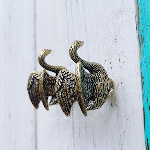 Könnte beinhalten: Ein bronzefarbener Ring mit einem Design aus stilisierten Schwanenflügeln. Der Ring hat eine strukturierte Oberfläche mit detaillierten Flügeln und Schwanenköpfen. Der Ring ist vor einem weißen und türkisfarbenen Hintergrund platziert.