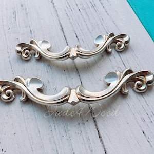 Puede incluir: Dos tiradores de cajón de plata adornados con un diseño de volutas decorativas. Los tiradores están sobre una superficie de madera blanca.