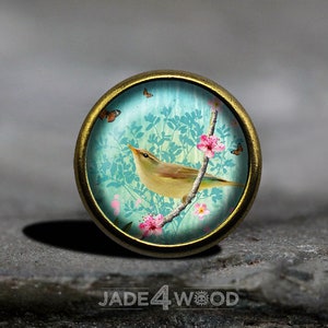 Könnte beinhalten: Ein runder Anhänger aus Messing mit einem Glaseinsatz, der ein blaues und grünes Blumenmuster mit einem kleinen Vogel zeigt, der auf einem Ast sitzt. Der Vogel ist gelb mit braunen Markierungen. Das Design enthält rosa Blumen und Schmetterlinge.