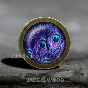 Könnte beinhalten: Ein runder Anhänger aus Bronze mit einem Glaseinsatz, der eine Nahaufnahme von Pfauenfedern in den Farben Lila, Blau und Türkis zeigt.