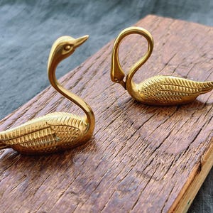 Könnte beinhalten: Zwei goldene Schwanenfiguren auf einer Holzoberfläche. Die Schwäne sind mit strukturierten Federn detailliert und haben ein realistisches Aussehen.