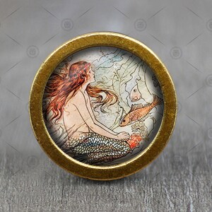 May include: A gold-toned round knob with a vintage illustration of a mermaid with red hair and a fish tail, surrounded by seaweed and fish. The knob has a raised edge.