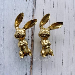 May include: Two gold-coloured rabbit-shaped cabinet knobs with a smiling face and arms crossed. The knobs are mounted on a white wooden surface.