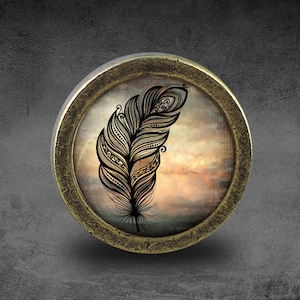 Può includere: Un pomello rotondo color ottone con un inserto in vetro che presenta un design di piuma stilizzato in bianco e nero su uno sfondo acquerellato rosa, arancione e giallo.