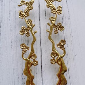 Op de afbeelding: Twee goudkleurige kastgrepen in een bloemtakontwerp. Elke greep heeft een gebogen, gestileerde tak met clusters van kleine bloemen. De grepen zijn gemaakt van een glanzend, metalen materiaal en staan tegen een witte houten achtergrond.