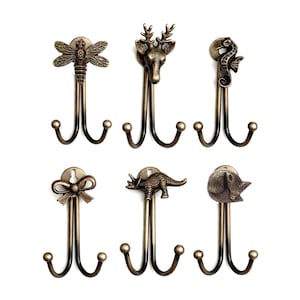 Pode incluir: Conjunto de seis ganchos de parede de metal cor bronze. Cada gancho apresenta um topo decorativo com diferentes desenhos, incluindo uma libélula, cabeça de veado, cavalo-marinho, laço, dinossauro e um peixe. Os ganchos têm um design de gancho duplo.