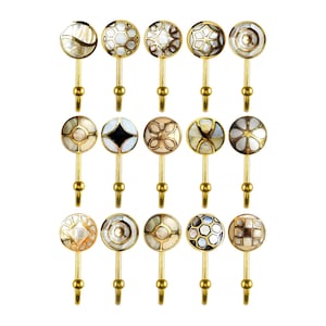 May include: Fifteen decorative brass hooks with various inlaid mother-of-pearl designs. The hooks have a gold finish and are arranged in three rows of five, showcasing a variety of geometric and floral patterns.