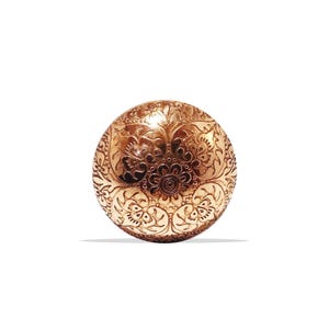 Könnte beinhalten: Ein detaillierter, goldfarbener Knopf mit einem aufwendigen Blumenmuster. Der runde Knopf weist ein erhabenes Design aus Blumen und Blättern auf, wodurch eine strukturierte Oberfläche entsteht. Die Gesamtästhetik ist verziert und dekorativ.