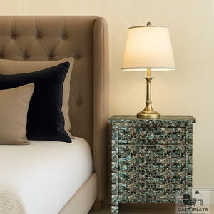 May include: A bedroom scene with a beige upholstered headboard and a patterned bedside table. A brass lamp with a white shade sits on the table. Beige and navy blue decorative pillows are against the headboard.