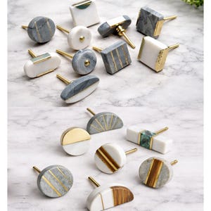 Puede incluir: Una colección de tiradores decorativos para armarios en varias formas y diseños. Los tiradores presentan detalles de mármol, oro y madera. Los estilos incluyen formas redondas, cuadradas y ovaladas con herrajes de latón. La paleta de colores incluye blanco, gris y marrón.