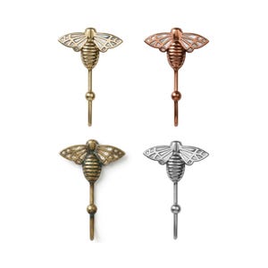 May include: Four decorative bee-shaped wall hooks in gold, copper, bronze, and silver finishes. Each hook features a detailed bee design with a hook extending from the bottom. The wings have a geometric pattern.