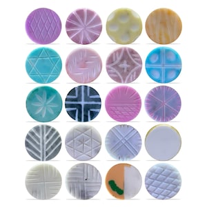 Könnte beinhalten: Eine Sammlung von einundzwanzig runden, dekorativen Knöpfen mit verschiedenen geschnitzten geometrischen und floralen Mustern. Die Knöpfe sind in den Farbtönen Lila, Rosa, Blau, Gelb und Weiß gehalten. Die Designs umfassen Diamanten, Sterne und kreuzartige Formen.