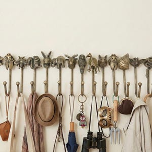 May include: A wall-mounted coat rack with bronze animal-shaped hooks. The hooks are holding items like keys, a hat, a scarf, an umbrella, binoculars, and a purse. Animals include a fox, a cicada, a bee, a squirrel, a rabbit, and an elephant. The rack is mounted on a cream-colored wall.