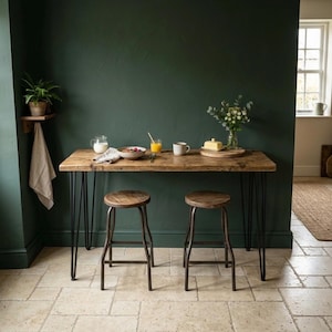 May include: A rectangular wooden table with a natural grain finish and four black metal hairpin legs, accompanied by two matching round wooden stools with black metal legs. The table surface holds a glass pitcher, a bowl, a glass, and a mug.