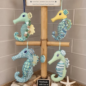 May include: Four handmade felt seahorse ornaments hanging from a wooden stand. The seahorses feature various shades of blue, green, and yellow with sequin and button embellishments. A sign at the top reads Handmade Felt Seahorses Coastal Charm Collection.