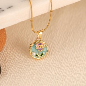 May include: A gold-colored necklace with a pendant featuring a floral design. The pendant has a light blue background with a pink and purple flower, a white pearl, and green leaves. The necklace is on a textured, off-white surface.