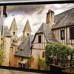 May include: Framed print of a European village scene. The image features stone buildings with dark, angled roofs, and a church with two tall spires. The sky is overcast with hints of blue. The print is in a black frame.