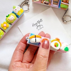 Peut inclure: Trois porte-clés rectangulaires ornés de breloques miniatures en 3D colorées. Un porte-clés présente des figurines de sushis et de boulettes de riz, un autre des tasses à café avec des motifs d'ours, et le troisième des motifs floraux et animaliers.
