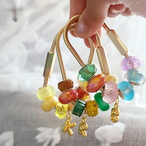 May include: Three gold-toned metal keychains featuring a variety of colourful, textured, and faceted beads in shades of yellow, orange, red, green, pink, and blue. Each keychain includes a small gold letter charm, specifically K and R.