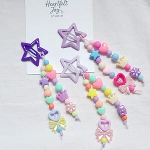 May include: Three star-shaped hair clips attached to a white display card labelled Heartfelt Joy STUDIO. Each clip features dangling strands of colourful plastic beads in shapes including hearts, stars, crowns, bows, and flowers in pastel tones.