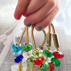 May include: A set of gold-toned metal carabiner-style keychains adorned with faceted, multi-coloured glass beads in shades of red, green, blue, and clear. Each keychain features a dangling gold-toned metal snowflake charm and a letter charm.