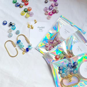 May include: A collection of colourful beads, gold-toned oval carabiner keychains, and gold letter charms. Some beads are loose, while others are packaged in iridescent holographic pouches with Heartfelt Joy Studio labels and branding.