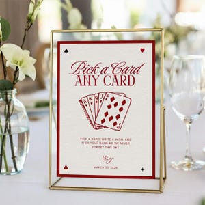Pode incluir: Um letreiro emoldurado com uma moldura de metal dourado e um cartão branco. O cartão tem texto vermelho que diz "Pick a Card ANY CARD" e apresenta gráficos de cartas de baralho. A parte inferior do cartão tem a data "MARÇO 30, 2026".
