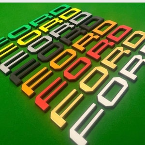 Puede incluir: Una colección de letras tridimensionales coloridas que deletrean la palabra "FORD" dispuestas sobre una superficie verde. Las letras son de varios colores, incluyendo verde, amarillo, negro, naranja, rojo y blanco.