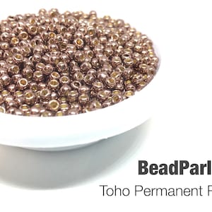 May include: A close-up of a bowl filled with small, round, metallic beads in a rose taupe color. The beads are Toho brand and are labeled "Toho Permanent Rose Taupe".