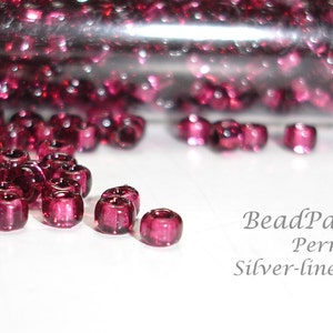 May include: Close-up of a small glass tube filled with cranberry red seed beads with a silver lining. The beads are scattered on a white surface. The text "BeadParlorBeads Permanent Silver-lined Cranberry" is visible below the beads.