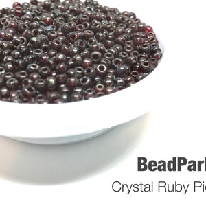 May include: A bowl filled with dark red glass beads with a silver sheen. The beads are small and round, and they are arranged in a random pattern. The bowl is white and has a smooth surface. The text "BeadParlorBeads Crystal Ruby Picasso 4504" is printed below the bowl.