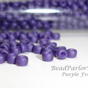 May include: Purple glass seed beads with a frosted finish. The beads are scattered on a white surface. BeadParlorBeads Purple Frost.
